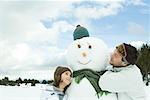 Brother and sister embracing snowman, smiling, head and shoulders
