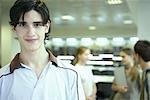 Male college student in library, smiling at camera, other students chatting in blurred background