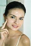 Young woman putting exfoliant mask on face, smiling at camera