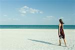 Woman walking across beach, carrying transparent container, full length, rear view