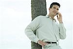 Man leaning against tree trunk, using cell phone, waist up