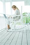 Woman sitting in rocking chair on porch, using laptop computer, full length