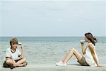 Garçon et sœur adolescente assis à côté de l'eau, parlant les uns aux autres par l'étain peut téléphoner