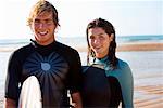 Couple standing with surfboard on beach smiling.
