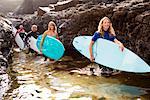 Four people carrying surfboards by large rocks smiling.