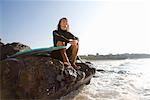 Woman sitting on large rocks with surfboard smiling.