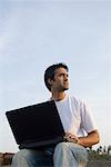 Low angle view of a young man holding a laptop on his lap and sitting at a coast, Madh Island, Mumbai, Maharashtra, India