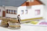 Businessman figurine sitting on stack of coins while reading, miniature house in background