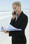 Businesswoman using cell phone and looking at file, standing on beach, smiling