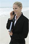 Businesswoman using cell phone, standing on beach, smiling