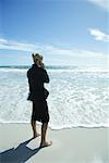 Businesswoman using cell phone, standing on beach barefoot, full length, side view