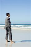 Businessman standing barefoot on beach with hands in pockets, side view