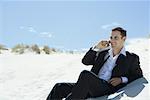 Businessman sitting on sand dune, using cell phone