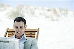 Businessman sitting in deck chair at beach, reading newspaper, smiling