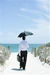 Businessman walking on sandy path leading to ocean, using umbrella, rear view