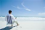 Man sitting in folding chair on beach, holding cell phone, rear view