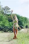 Young woman standing in rural field with arm raised, looking up, apple in hand