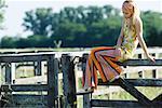 Young woman sitting on top of rural fence, looking down