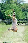 Young woman jumping with arms raised in rural setting, smiling at camera