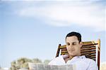 Young man sitting in deck chair, reading newspaper