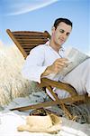 Young man sitting in deck chair on beach, reading newspaper