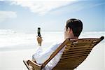 Man sitting in chair on beach, using cell phone, rear view