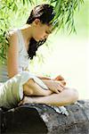 Teenage girl sitting on log under tree, looking down at foot