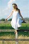 Young woman in dress sitting on rural fence with arms out, eyes closed