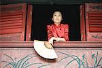 Young woman wearing traditional Chinese clothing, holding fan, leaning on windowsill
