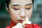 Young woman wearing traditional Chinese clothing, holding flower up to face, eyes closed, portrait