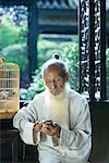 Elderly man wearing traditional Chinese clothing, using cell phone, next to bird cage