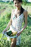 Young woman standing in garden, holding fresh produce in dress, smiling at camera