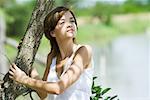 Young woman holding onto tree, looking away, waist up