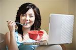 Young woman playing with laptop, eating with spoon