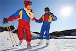Jeune couple tenant des skis et bâtons de ski avec une pente de ski à l'arrière-plan, souriant