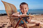 Boy sitting on chair at beach, smiling, portrait