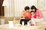 Grandmother, Mother and daughter reading in sofa in front of food at home