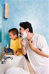 Side profile of a mid adult man shaving with his son sitting beside him in the bathroom