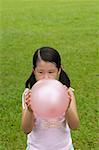 Girl standing on grass, blowing up pink balloon