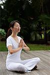 Woman sitting on porch, in tropical setting.  legs crossed, hands in namaste, yoga posture. eyes closed