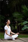 Woman in tropical setting, meditating on porch, eyes closed, in yoga OM posture, side view.
