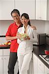 Man and woman in kitchen holding salad bowl and looking at camera smiling