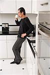 Man in kitchen, leaning on counter, drinking coffee, side profile to camera.