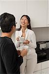 Femme assise sur le comptoir dans la cuisine rire avec l'homme face à son et son retour vers la caméra. les deux tasses de café holding.