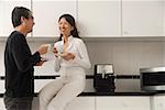 Woman sitting on counter in kitchen with man standing next to her, looking at each other, holding coffee cups and smiling