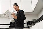 Man in kitchen, holding coffee cup and newspaper and looking at camera