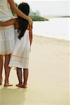 Mother and daughter standing on beach, arms around each other, rear view