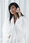 woman in bathrobe, standing in front of window, talking on phone