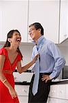 Man and woman in kitchen, woman laughing and tying mans tie.  Man with hands on hip.