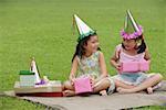 Two girls wearing party hats sitting on picnic blanket, holding pink gift boxes